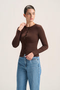 A woman modeling a fitted, long-sleeve brown button-front top paired with light blue jeans, standing against a plain light beige studio background.