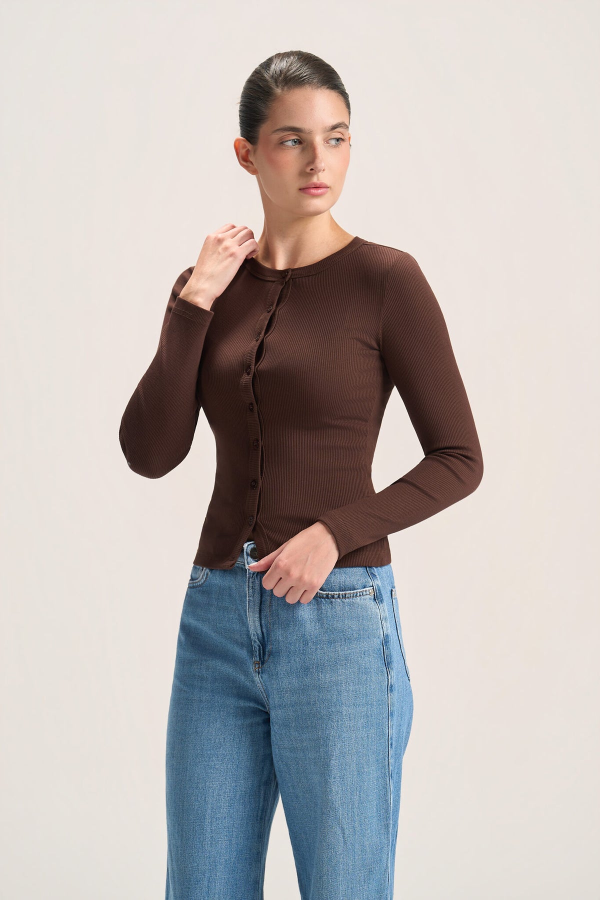 A woman modeling a fitted, long-sleeve brown button-front top paired with light blue jeans, standing against a plain light beige studio background.