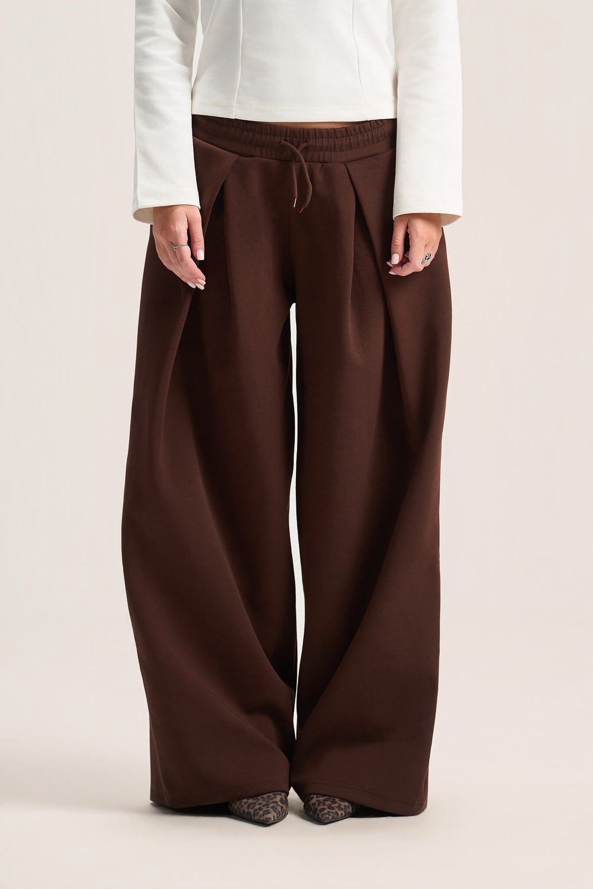 A person wearing wide-leg brown drawstring pants with a white long-sleeve top, shown from the chest down against a plain light beige studio background.