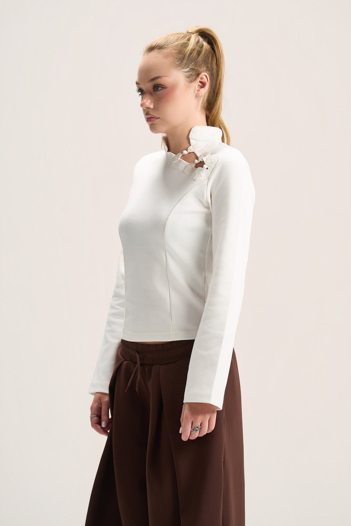 A woman wearing a fitted white long-sleeve mock-neck top with a decorative collar and dark brown wide-leg pants, photographed against a plain light background.