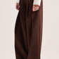A model wearing a pair of wide-leg brown trousers with a drawstring waist, paired with a white long-sleeve top and patterned flats, against a plain light beige studio background.
