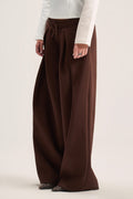 A model wearing a pair of wide-leg brown trousers with a drawstring waist, paired with a white long-sleeve top and patterned flats, against a plain light beige studio background.
