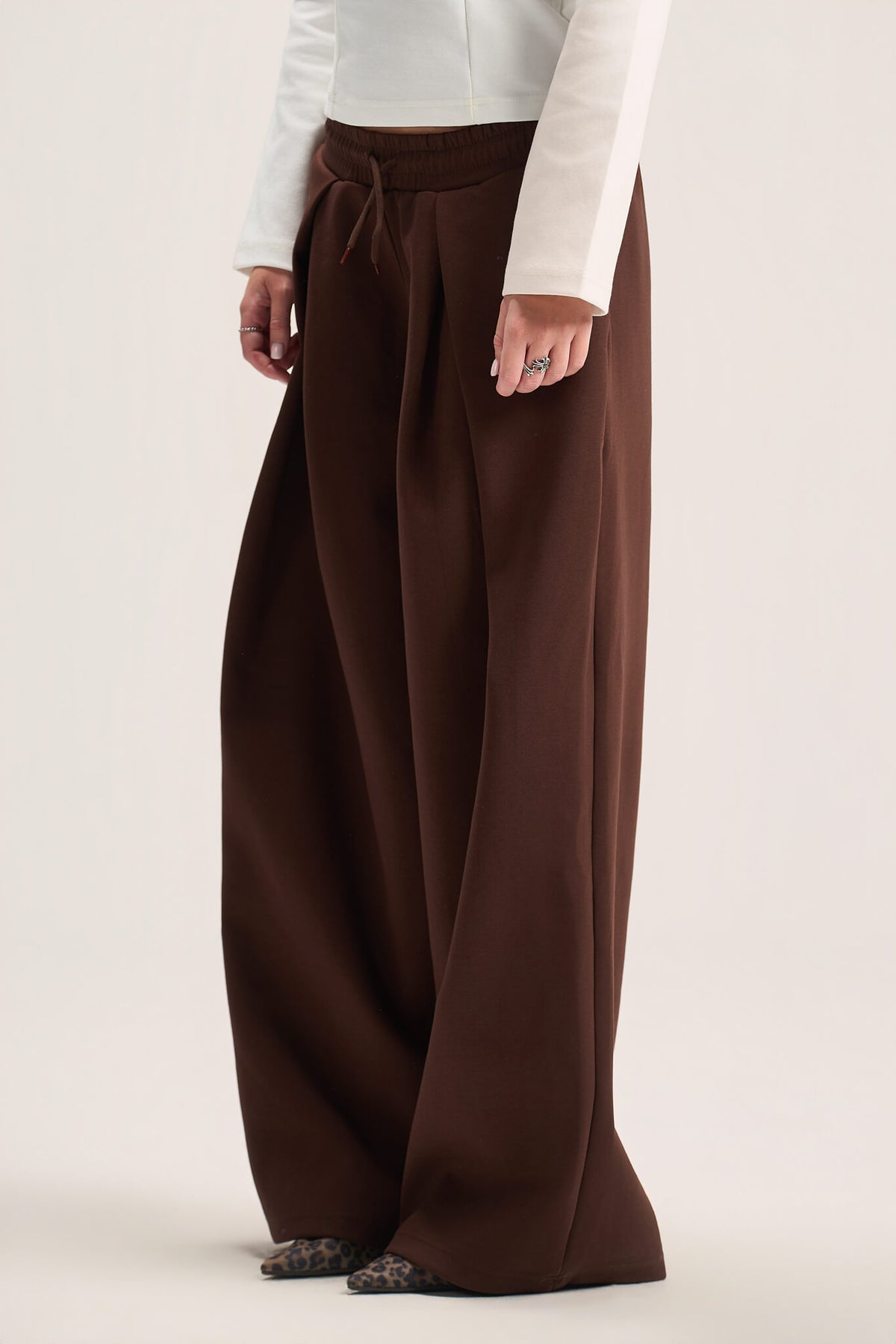 A model wearing a pair of wide-leg brown trousers with a drawstring waist, paired with a white long-sleeve top and patterned flats, against a plain light beige studio background.