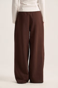 A person wearing loose, wide-leg brown pants with a white long-sleeve top, shown from the back against a plain light beige studio background.