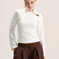 A woman modeling a fitted white long-sleeve top with a high mock neck and a small brown leather accent near the collar, paired with dark brown drawstring trousers, against a plain light beige studio background.