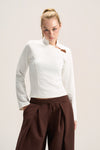 A woman modeling a fitted white long-sleeve top with a high mock neck and a small brown leather accent near the collar, paired with dark brown drawstring trousers, against a plain light beige studio background.