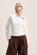 A woman modeling a fitted white long-sleeve top with a high mock neck and a small brown leather accent near the collar, paired with dark brown drawstring trousers, against a plain light beige studio background.