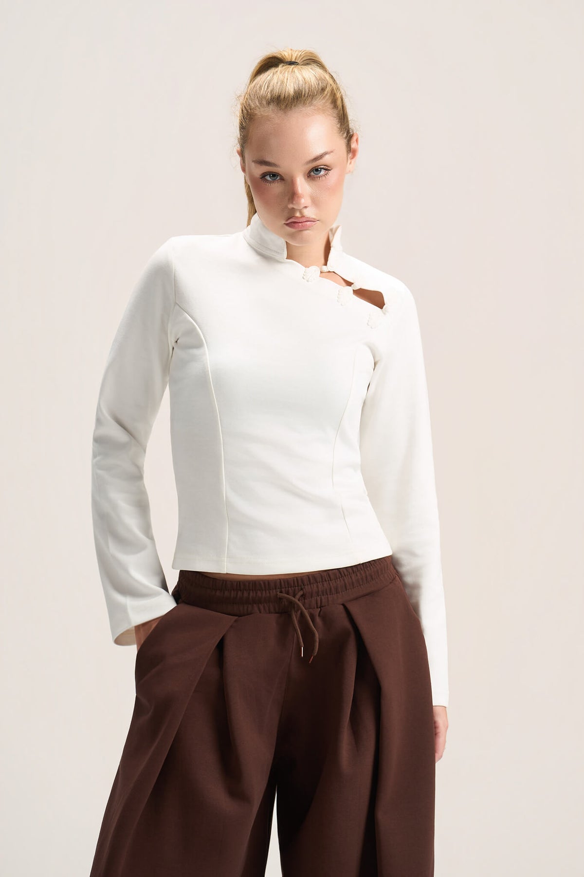 A woman modeling a fitted white long-sleeve top with a high mock neck and a small brown leather accent near the collar, paired with dark brown drawstring trousers, against a plain light beige studio background.