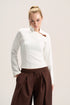 A woman modeling a fitted white long-sleeve top with a high mock neck and a small brown leather accent near the collar, paired with dark brown drawstring trousers, against a plain light beige studio background.