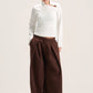A model stands against a plain light beige studio background wearing a white long-sleeve mock-neck top with a small brown accent on the chest and very wide-leg dark brown pants.