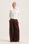 A model stands against a plain light beige studio background wearing a white long-sleeve mock-neck top with a small brown accent on the chest and very wide-leg dark brown pants.