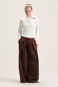 A model stands against a plain light beige studio background wearing a white long-sleeve mock-neck top with a small brown accent on the chest and very wide-leg dark brown pants.