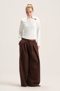 A model stands against a plain light beige studio background wearing a white long-sleeve mock-neck top with a small brown accent on the chest and very wide-leg dark brown pants.