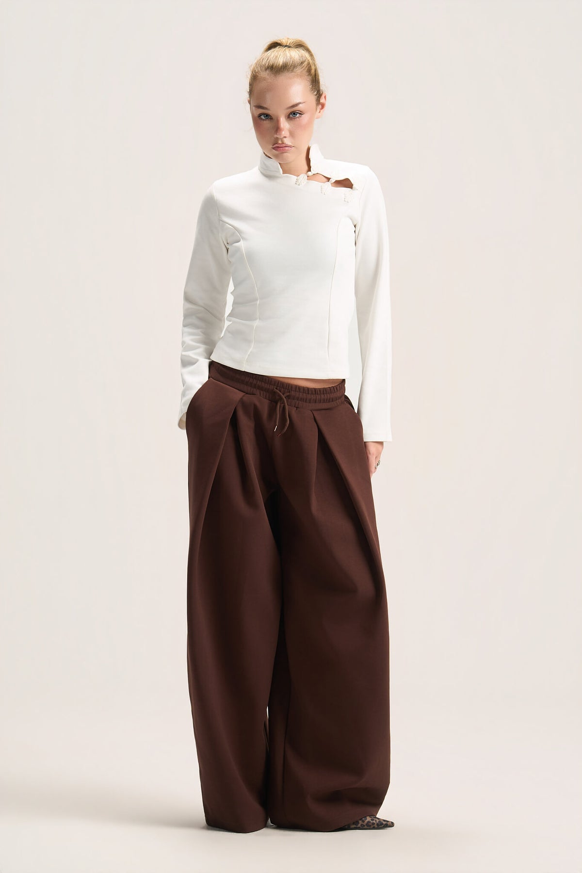 A model stands against a plain light beige studio background wearing a white long-sleeve mock-neck top with a small brown accent on the chest and very wide-leg dark brown pants.