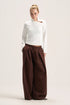 A model stands against a plain light beige studio background wearing a white long-sleeve mock-neck top with a small brown accent on the chest and very wide-leg dark brown pants.