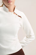 A close-up of a model wearing a white long-sleeve top with a mock neck and decorative cutout details on the chest, paired with brown bottoms against a light beige background.