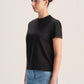 A woman wearing a black short-sleeve crewneck t-shirt and light blue jeans, standing against a plain light gray studio background.