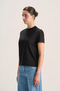 A woman wearing a black short-sleeve crewneck t-shirt and light blue jeans, standing against a plain light gray studio background.