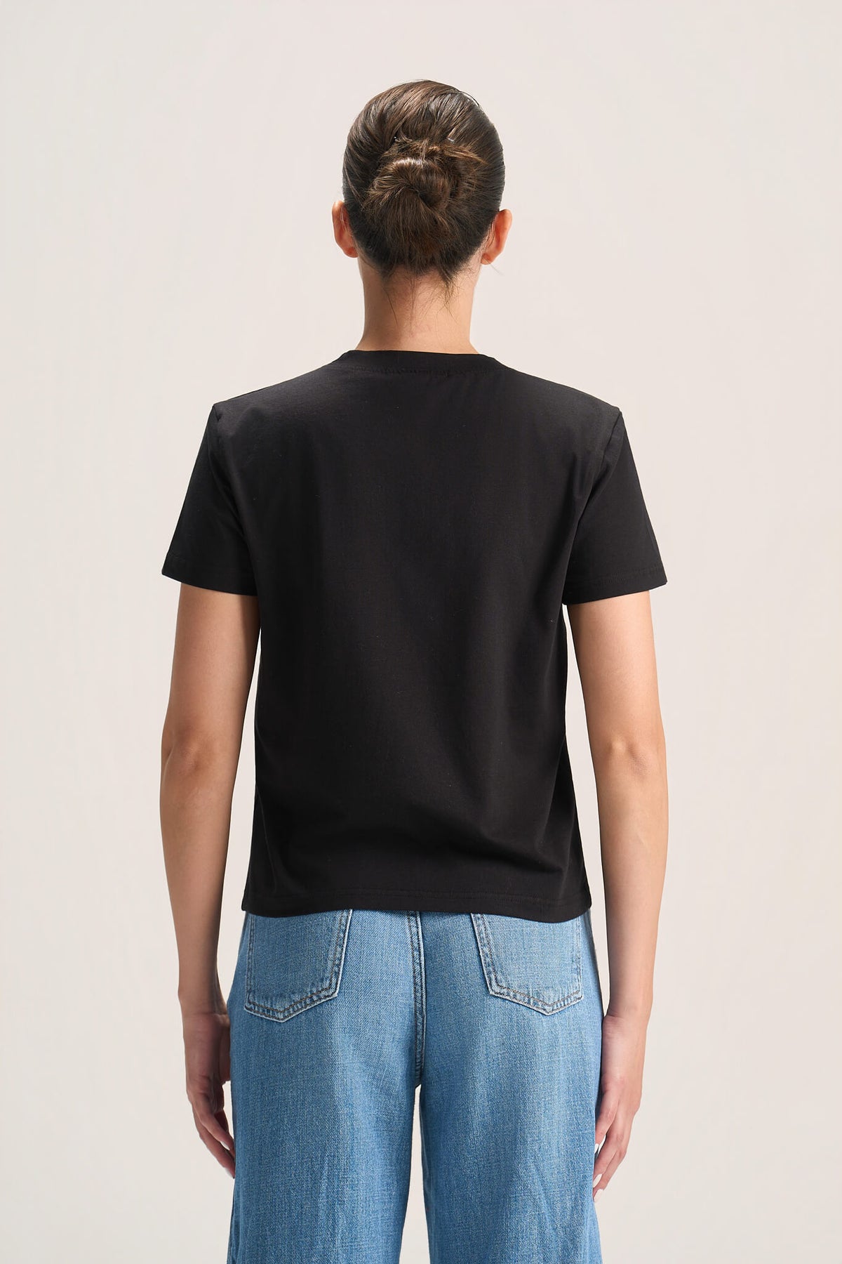 A woman viewed from the back wearing a plain black short-sleeve crew-neck T-shirt and light blue jeans against a light beige studio background.