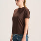 A woman with blonde hair in a ponytail wearing a short-sleeve brown mock neck t-shirt and blue jeans, standing against a light gray studio background.