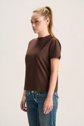 A woman with blonde hair in a ponytail wearing a short-sleeve brown mock neck t-shirt and blue jeans, standing against a light gray studio background.