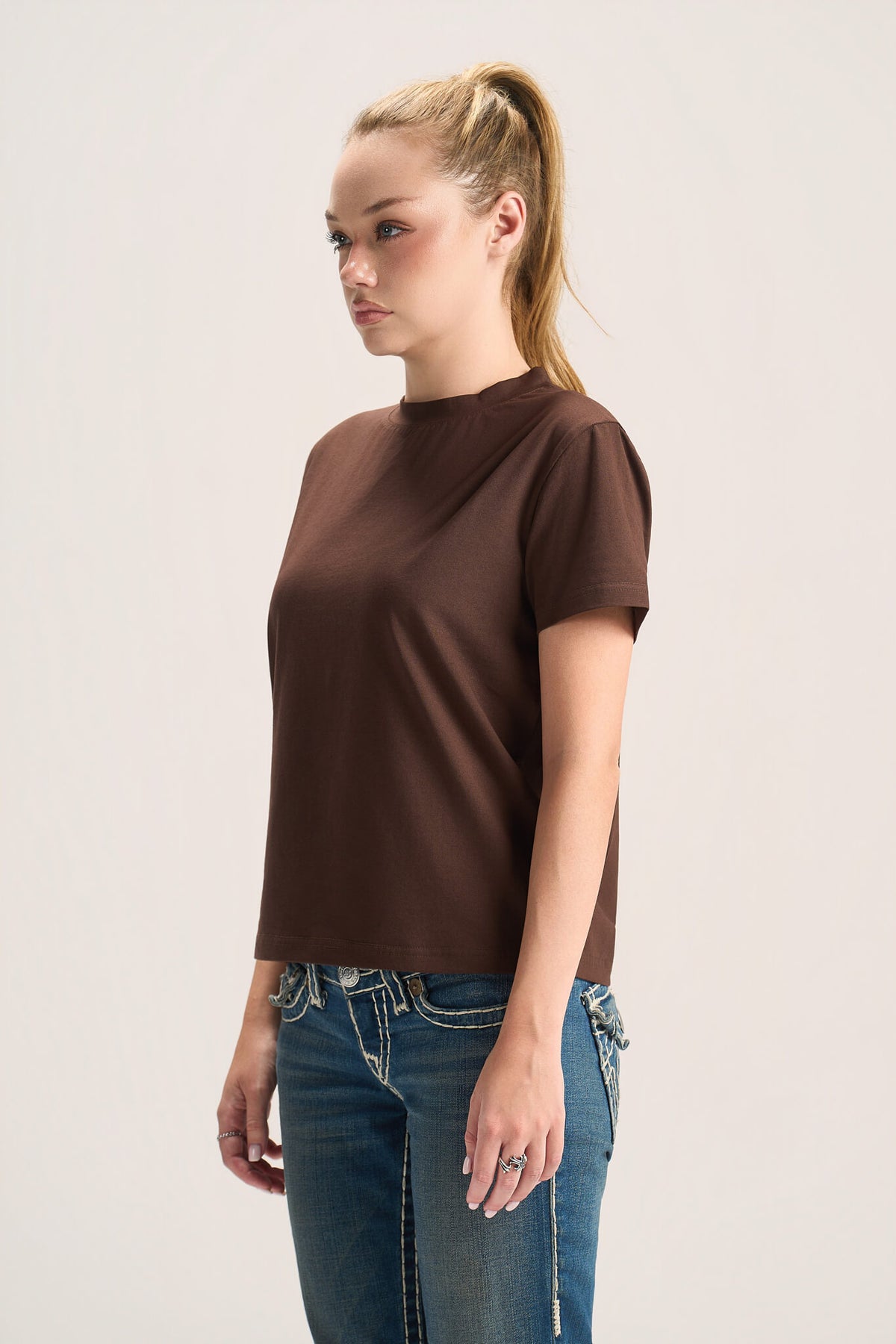 A woman with blonde hair in a ponytail wearing a short-sleeve brown mock neck t-shirt and blue jeans, standing against a light gray studio background.