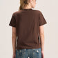A woman viewed from the back wearing a plain brown short-sleeve t-shirt and blue jeans with contrast stitching, set against a light gray studio background.