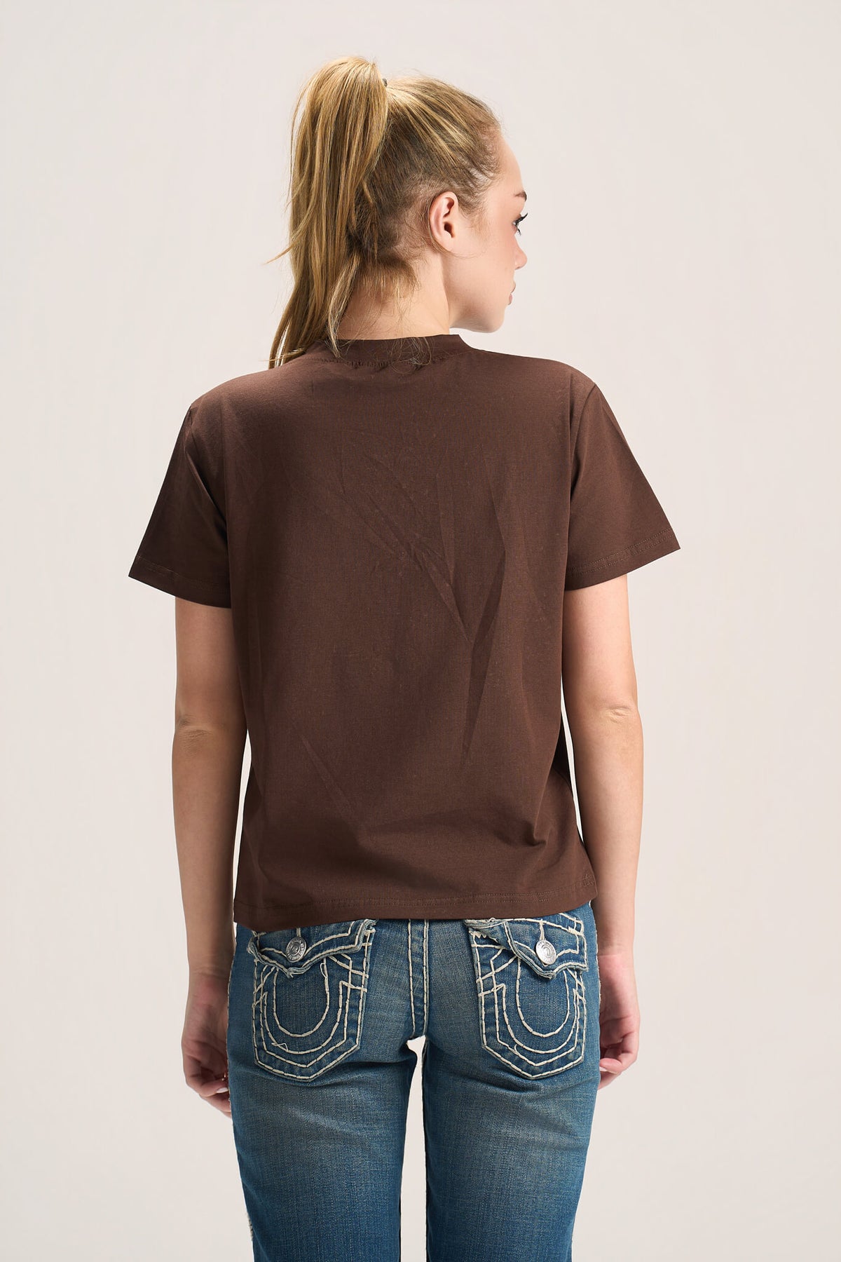 A woman viewed from the back wearing a plain brown short-sleeve t-shirt and blue jeans with contrast stitching, set against a light gray studio background.