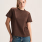 A woman modeling a brown short-sleeve crewneck t-shirt paired with blue jeans, photographed against a plain light gray studio background.