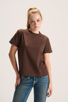 A woman modeling a brown short-sleeve crewneck t-shirt paired with blue jeans, photographed against a plain light gray studio background.
