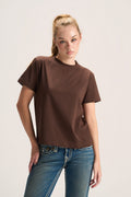 A woman modeling a brown short-sleeve crewneck t-shirt paired with blue jeans, photographed against a plain light gray studio background.