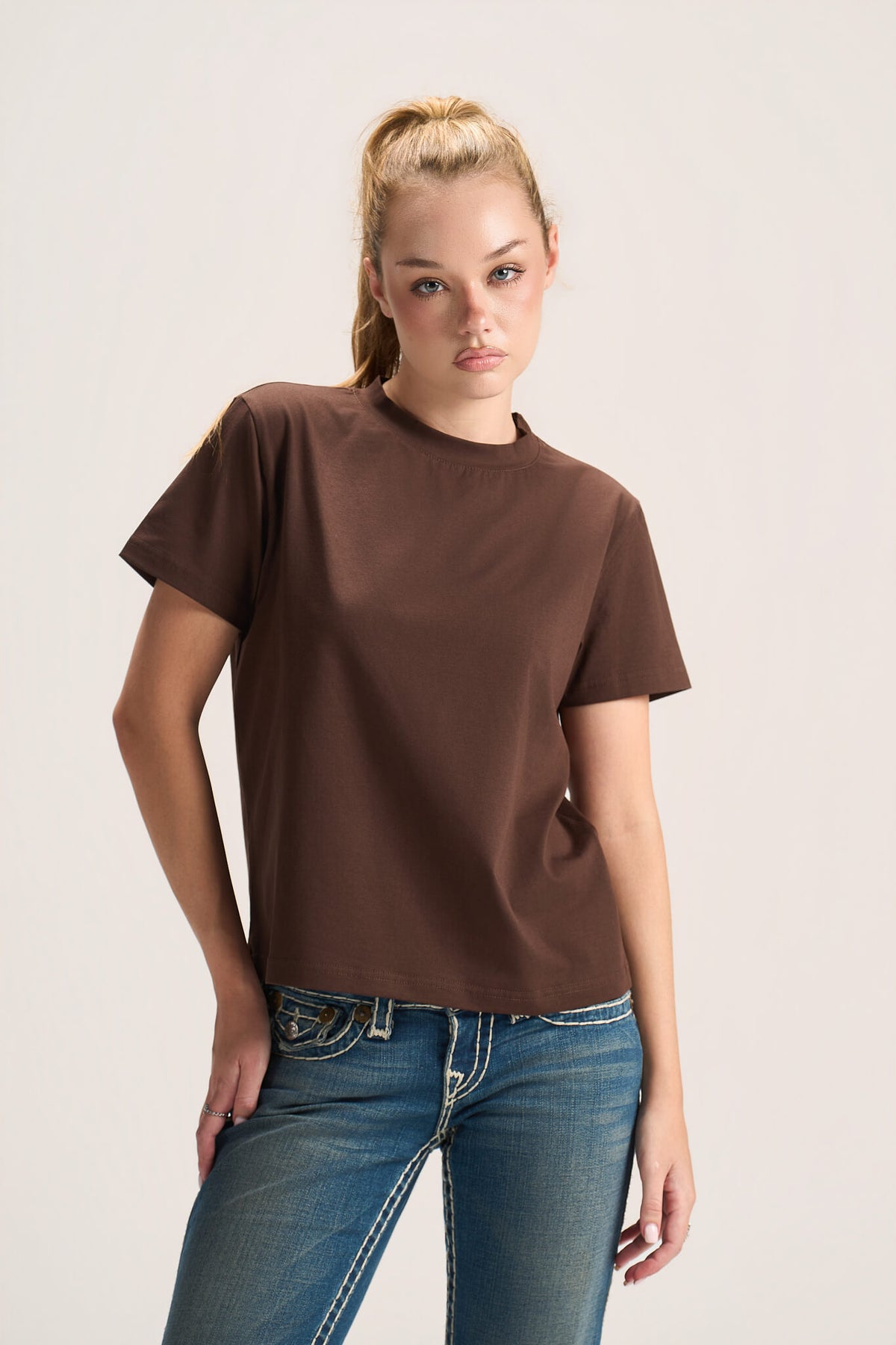 A woman modeling a brown short-sleeve crewneck t-shirt paired with blue jeans, photographed against a plain light gray studio background.