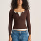 A model with curly dark hair wearing a brown ribbed long-sleeve henley top over a white undershirt and blue jeans, standing against a light beige studio background.