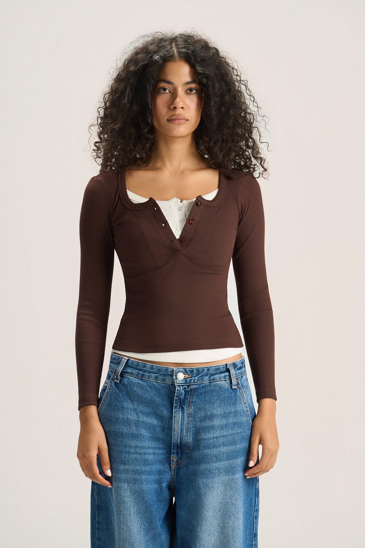 A model with curly dark hair wearing a brown ribbed long-sleeve henley top over a white undershirt and blue jeans, standing against a light beige studio background.
