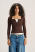 A model with curly dark hair wearing a brown ribbed long-sleeve henley top over a white undershirt and blue jeans, standing against a light beige studio background.