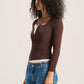 A woman with curly dark hair is shown in profile wearing a brown long-sleeve henley top over a white undershirt and blue jeans, photographed against a plain light background.