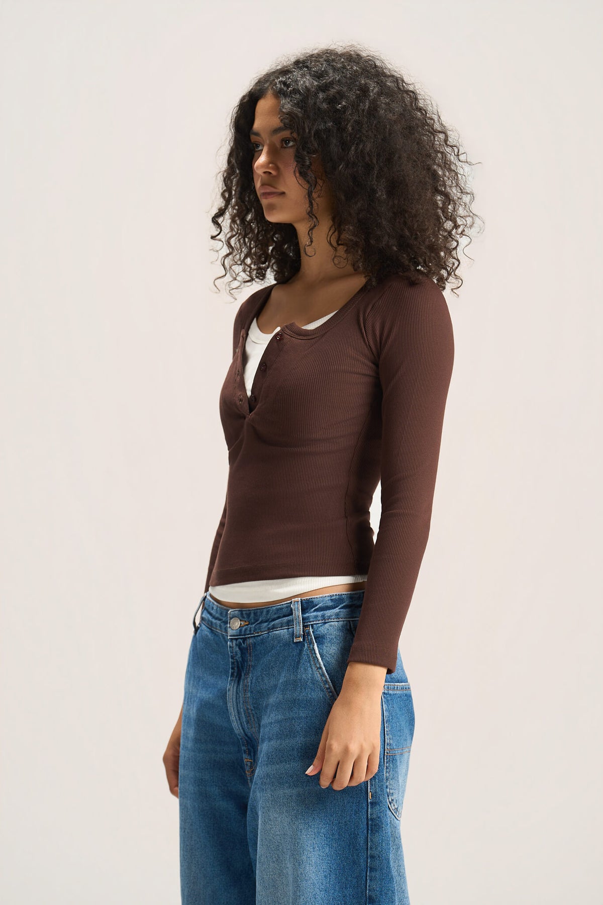 A woman with curly dark hair is shown in profile wearing a brown long-sleeve henley top over a white undershirt and blue jeans, photographed against a plain light background.