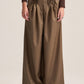 A model wearing high-waisted, wide-leg brown trousers with a gathered elastic waistband, paired with a dark brown long-sleeve top and dark brown shoes, against a solid light beige background.