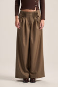 A model wearing high-waisted, wide-leg brown trousers with a gathered elastic waistband, paired with a dark brown long-sleeve top and dark brown shoes, against a solid light beige background.