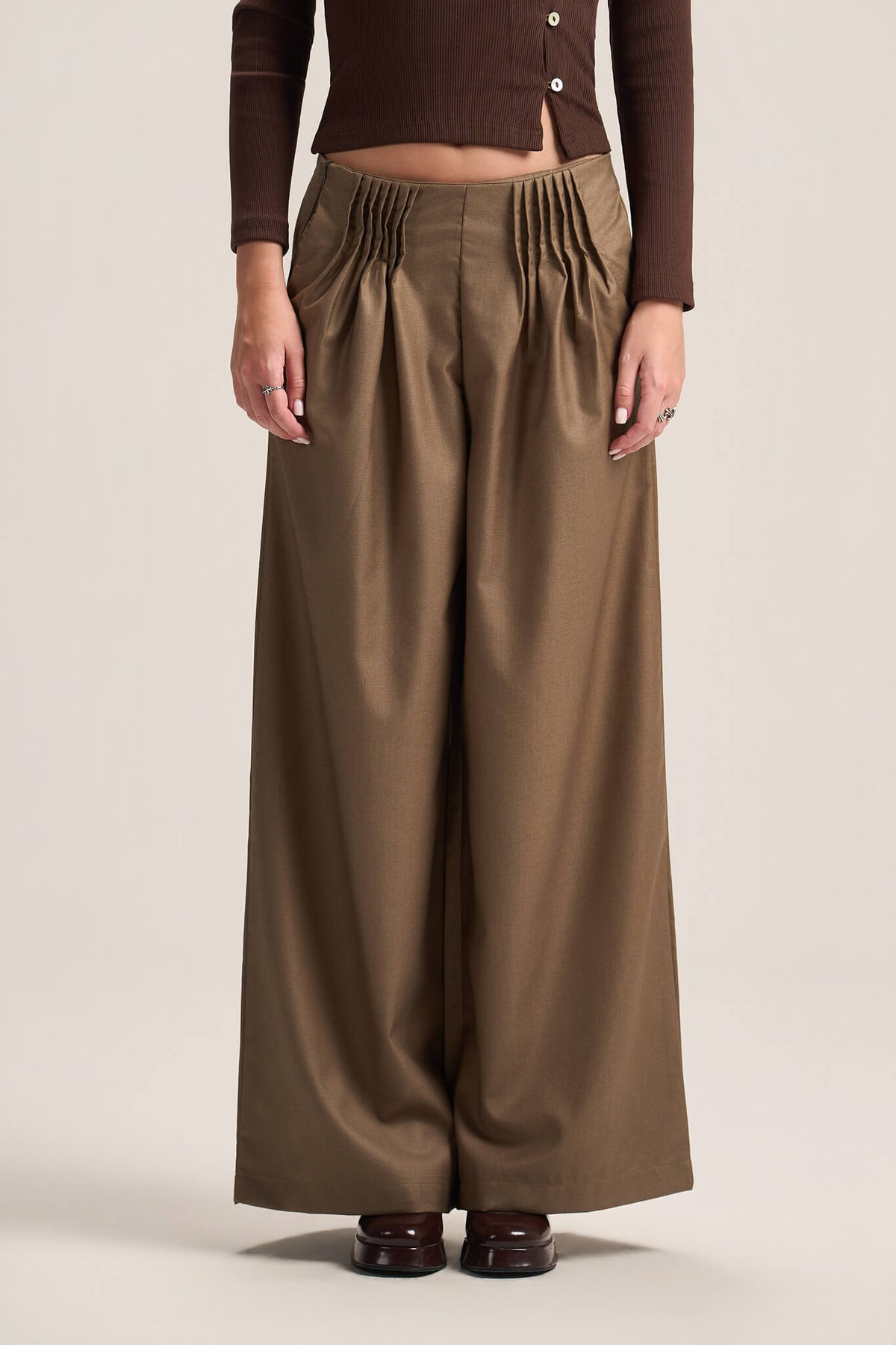 A model wearing high-waisted, wide-leg brown trousers with a gathered elastic waistband, paired with a dark brown long-sleeve top and dark brown shoes, against a solid light beige background.