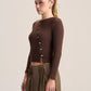 A woman modeling a dark brown ribbed long-sleeve crop top with a button-down front, paired with olive green pleated trousers, against a plain light beige studio background.