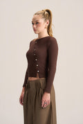 A woman modeling a dark brown ribbed long-sleeve crop top with a button-down front, paired with olive green pleated trousers, against a plain light beige studio background.