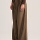 A model wearing high-waisted, wide-leg pleated trousers in a warm brownish-olive shade, paired with a dark brown cropped cardigan and dark brown platform shoes, against a solid light beige studio background.