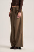 A model wearing high-waisted, wide-leg pleated trousers in a warm brownish-olive shade, paired with a dark brown cropped cardigan and dark brown platform shoes, against a solid light beige studio background.