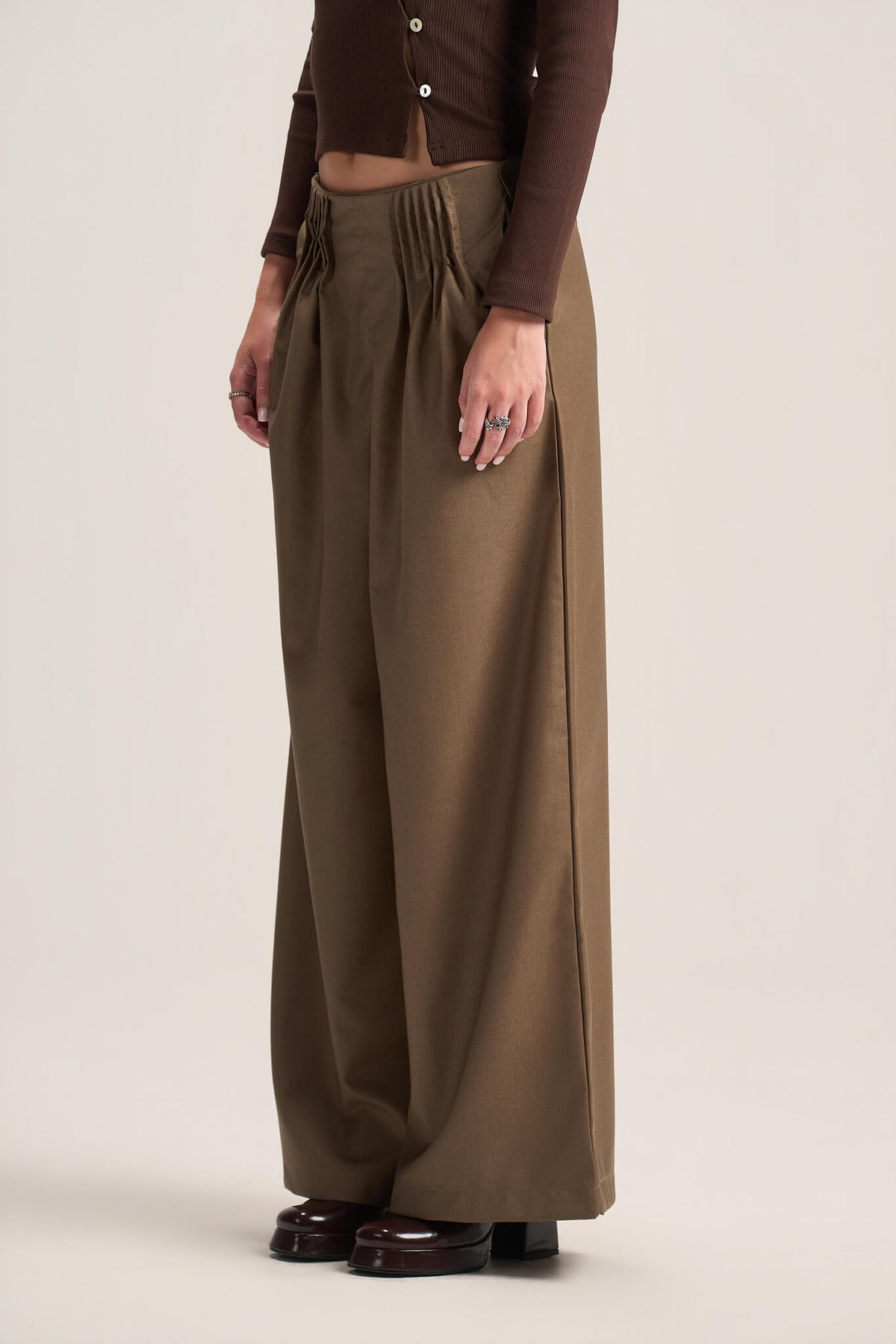 A model wearing high-waisted, wide-leg pleated trousers in a warm brownish-olive shade, paired with a dark brown cropped cardigan and dark brown platform shoes, against a solid light beige studio background.