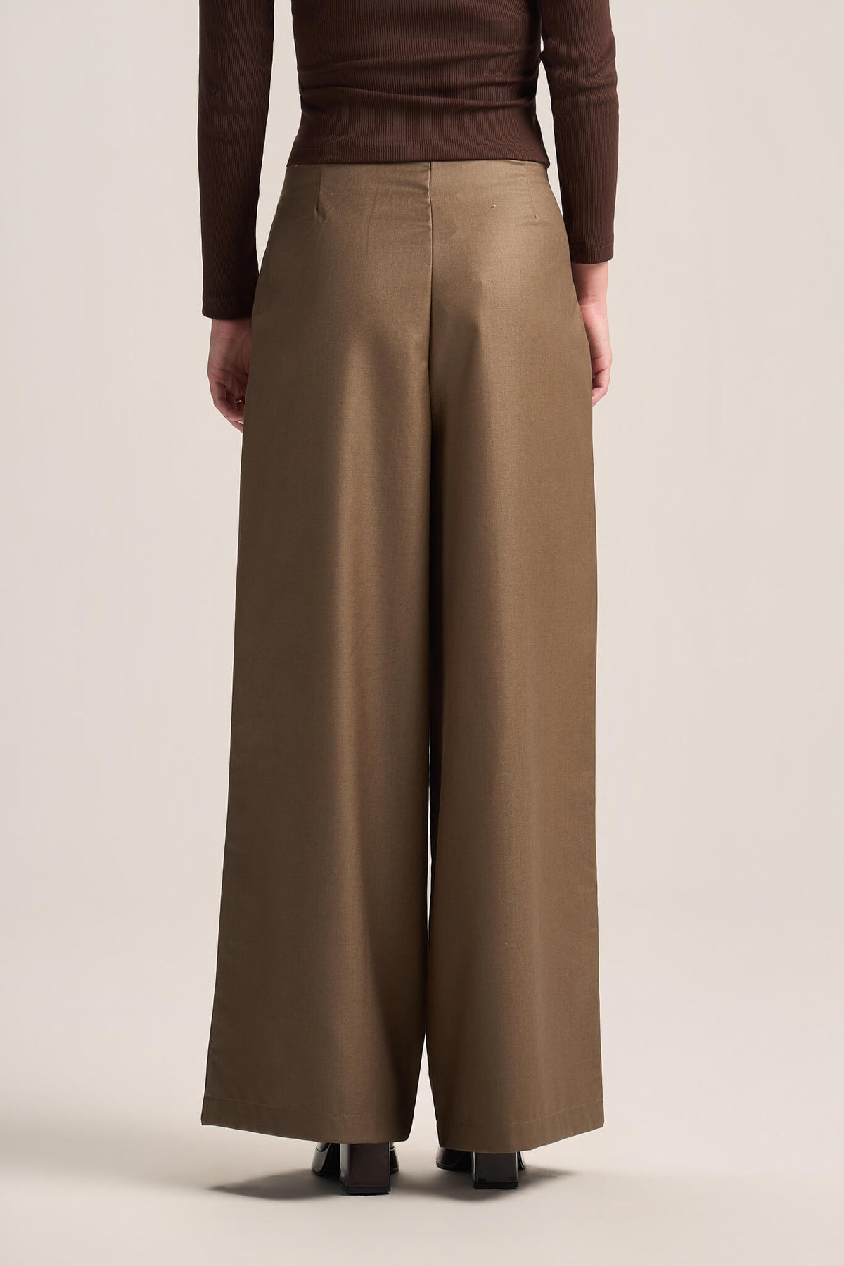 A model shown from the back wearing high-waisted, wide-leg trousers in a warm taupe-brown color, paired with a dark brown long-sleeve top and black shoes, against a solid light beige studio background.