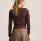 A woman shown from the back wearing a fitted long-sleeve brown top and wide-leg taupe pants against a plain light beige studio background.