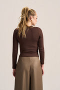 A woman shown from the back wearing a fitted long-sleeve brown top and wide-leg taupe pants against a plain light beige studio background.