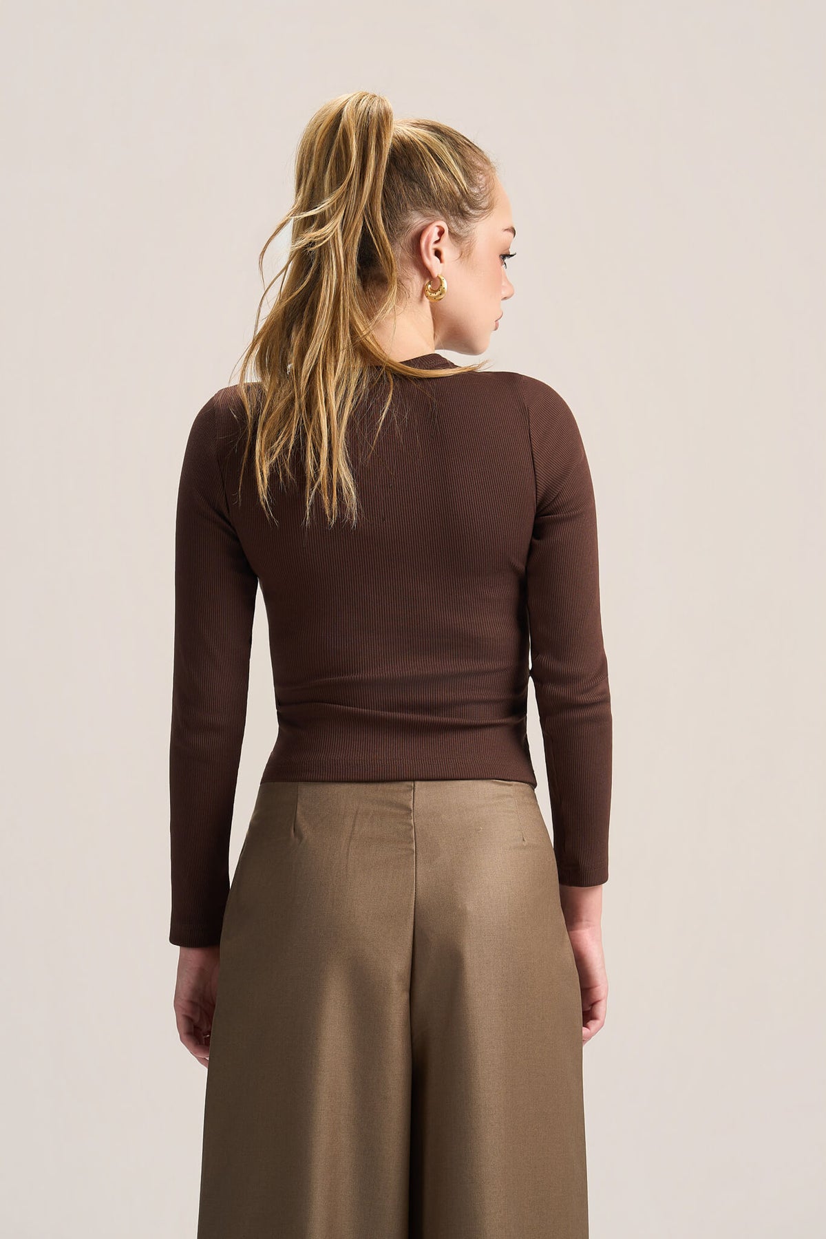 A woman shown from the back wearing a fitted long-sleeve brown top and wide-leg taupe pants against a plain light beige studio background.