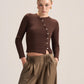 A model wearing a brown long-sleeve button-front crop top and high-waisted brown pleated trousers, standing against a light beige studio background.